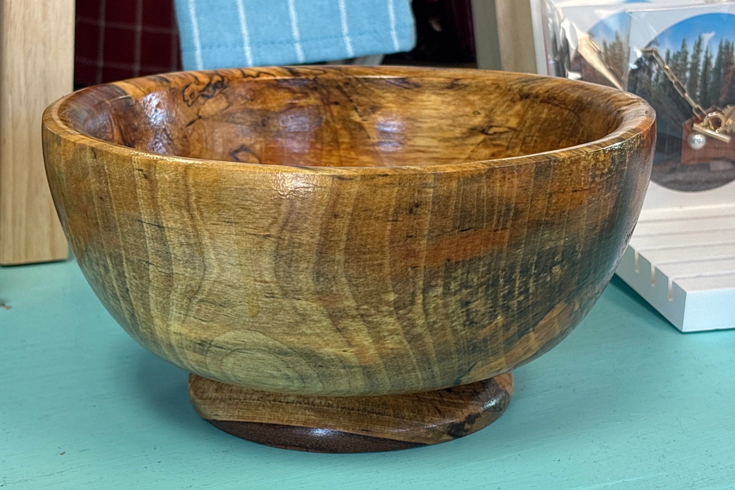 A handcrafted wooden bowl with a glossy finish and natural grain patterns, sitting on a turquoise painted wooden table with a rustic background.