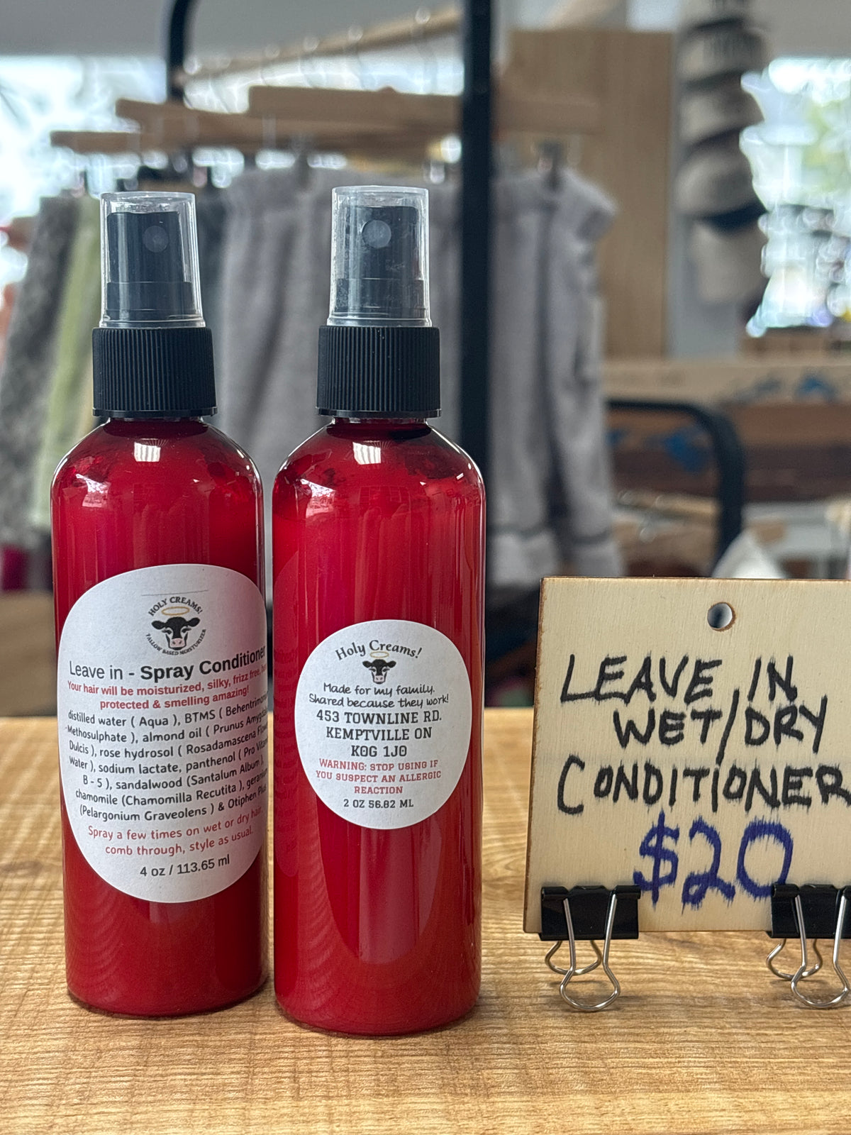 Two red plastic spray bottles of hair care products on a wooden table, next to a handwritten price sign that reads 'LEAVE IN WET/DRY CONDITIONER $20.00'.
