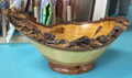 A handcrafted wooden bowl with a natural bark edge and a footed base, displayed on a turquoise surface with a blurred background of hanging colorful items.