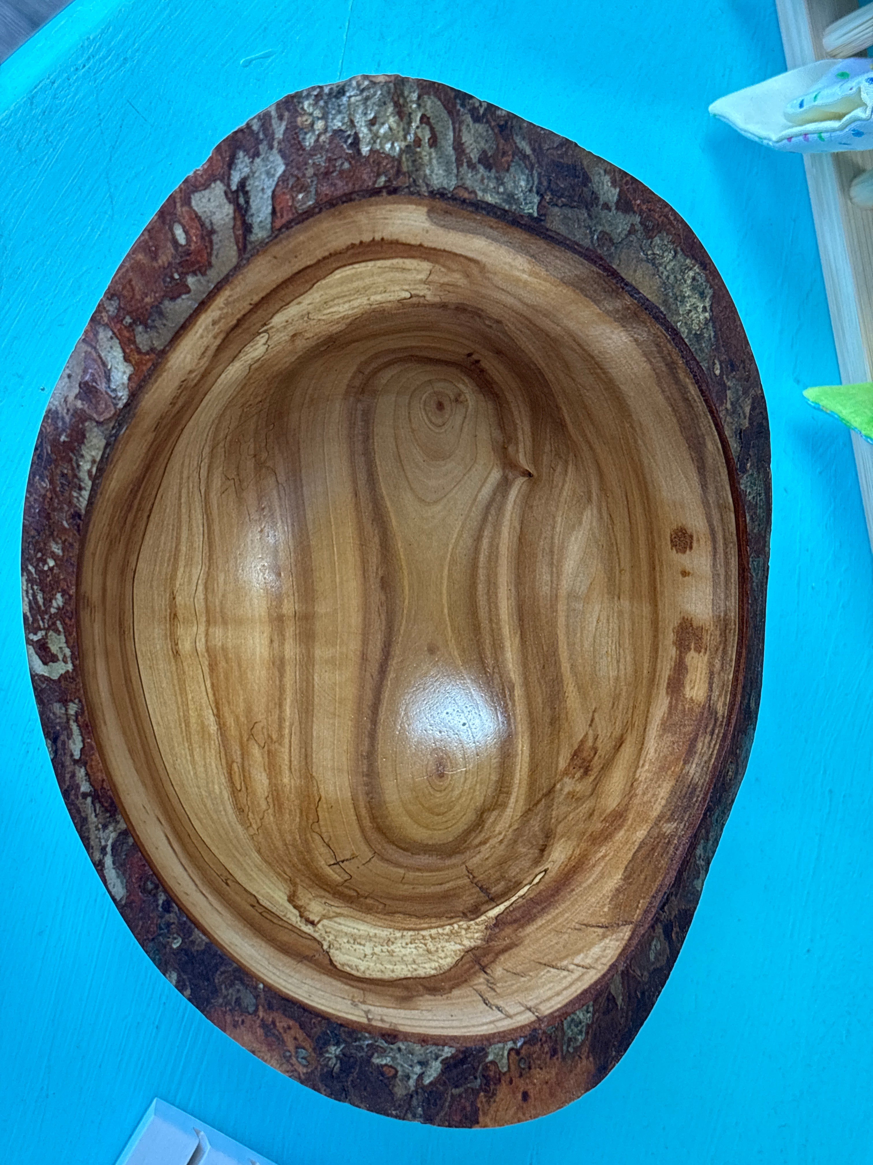 A rustic handcrafted wooden bowl with a natural bark edge and polished light brown interior, photographed from above on a bright blue surface.