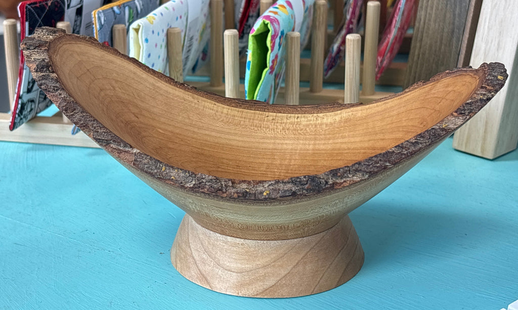A handmade wooden bowl with a natural bark edge and a carved wooden stand, displayed on a turquoise table in front of a fabric display stand.