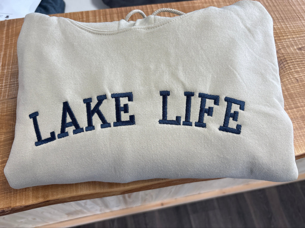 folded sand coloured hoodie with 'Lake Life' embroidery on a wooden surface