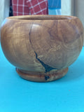 A round wooden bowl with a natural wood grain pattern and a prominent dark fissure running down the front, sitting on a bright blue surface.