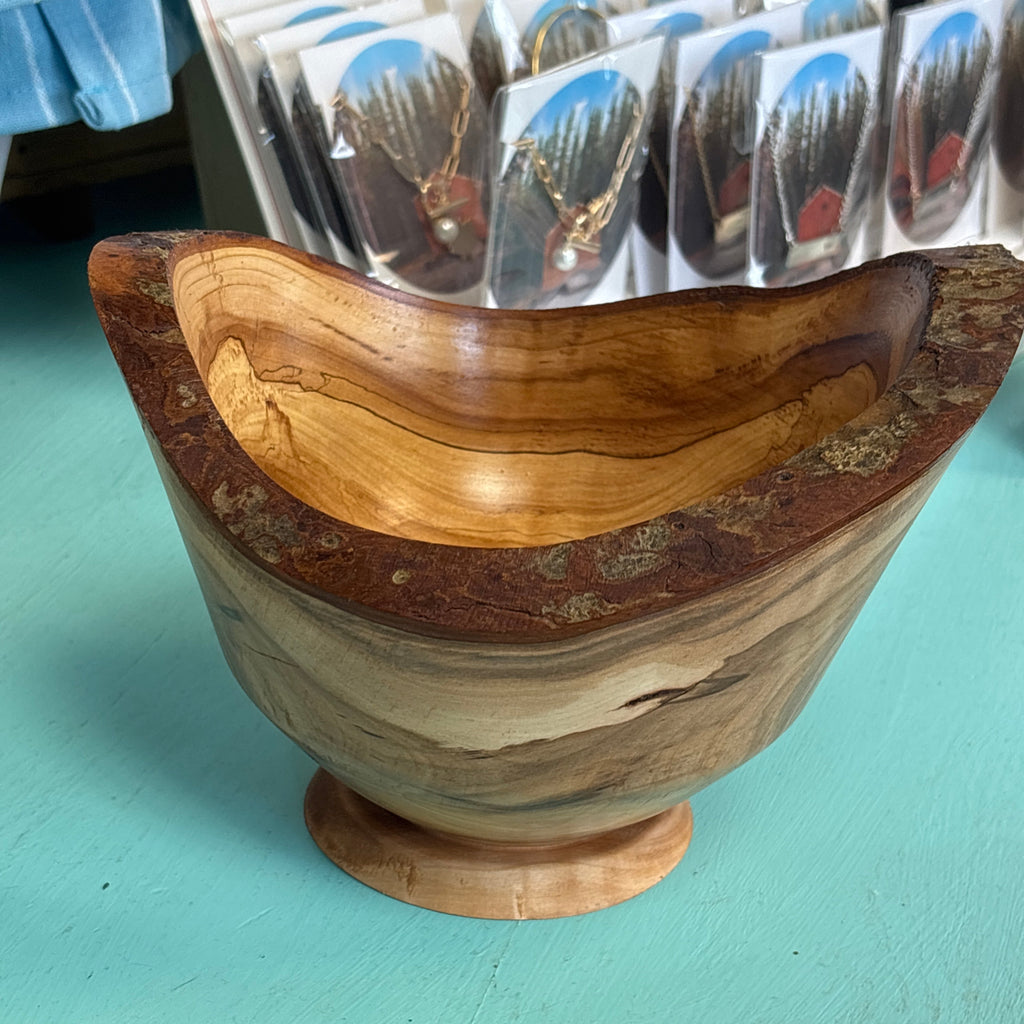 A handcrafted wooden bowl with a rustic live edge and a small pedestal base, displayed on a turquoise surface with packaged jewelry in the background.