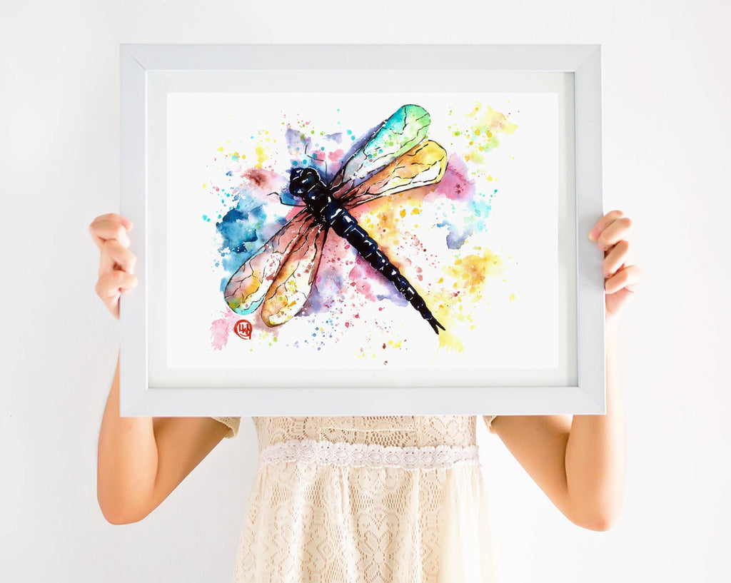 Person holding a framed watercolor painting of a dragonfly with colorful splashes on a white background