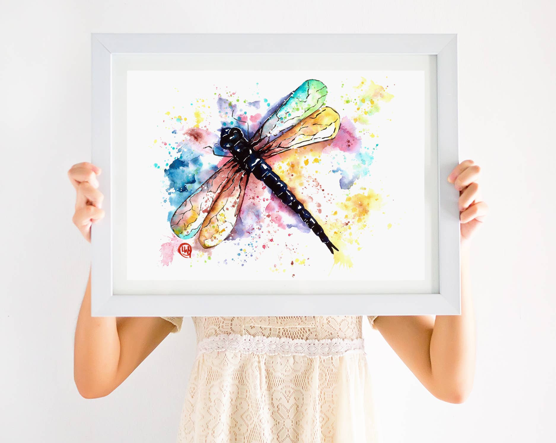 Person holding a framed watercolor painting of a dragonfly with colorful splashes on a white background
