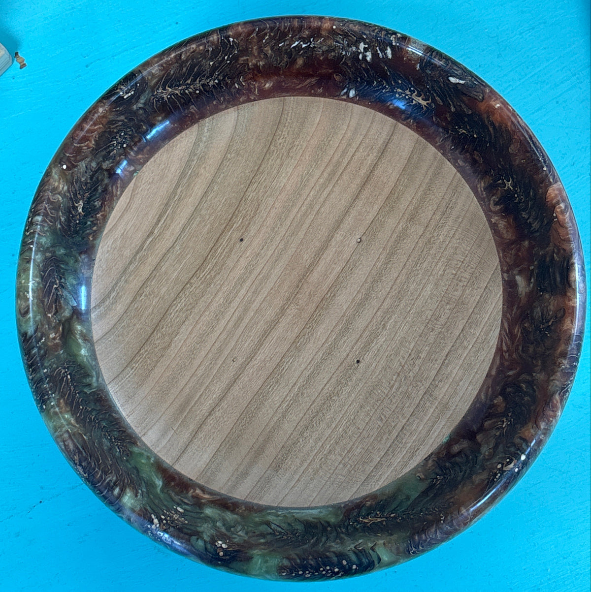 A round wooden bowl with a polished light brown wood-grain center and a thick outer rim featuring a marbled pattern of dark brown, black, and green, shown from a top-down perspective on a bright blue background.