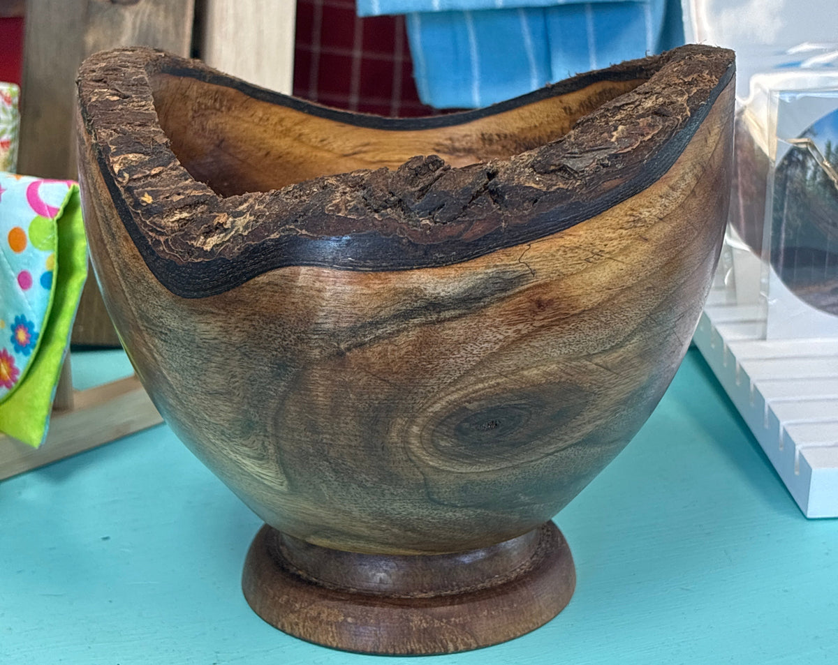 A rustic handcrafted wooden bowl with a live edge and natural bark, displayed on a turquoise surface with a blue striped towel in the background.