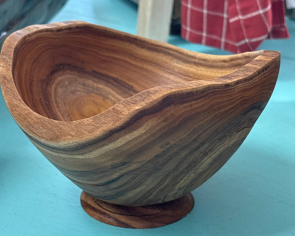 A handcrafted wooden bowl with a wavy, organic rim and a small round pedestal base, made from wood with prominent wavy grain patterns in brown and tan tones, set on a turquoise surface.