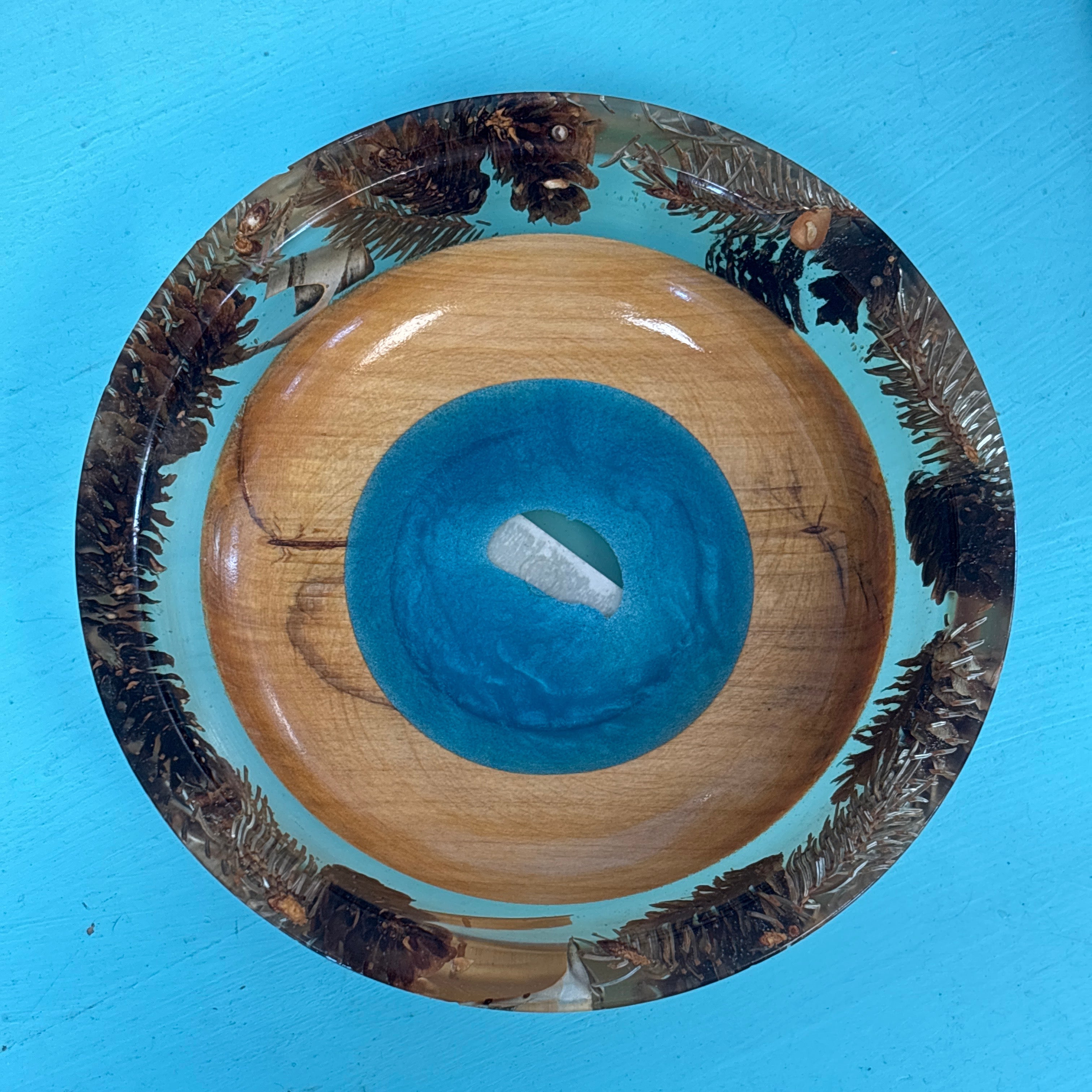 A round wooden bowl with a natural wood grain center, a bright turquoise blue circular inlay, and a dark brown pine-cone patterned rim, shown from a top-down view on a bright blue background.