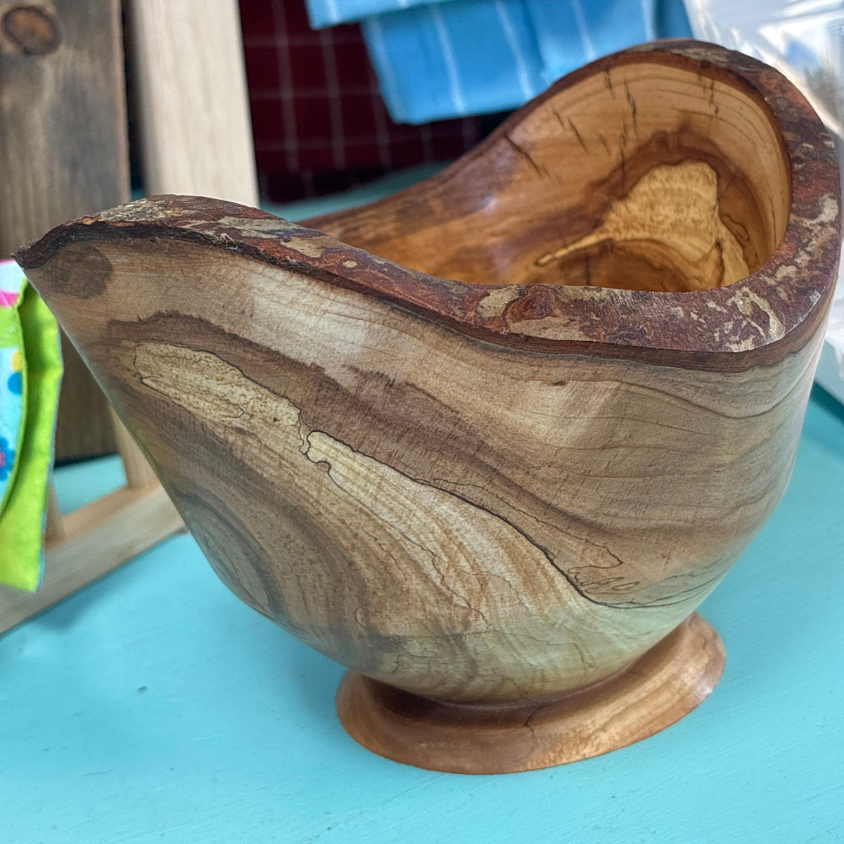A handcrafted wooden bowl with a natural organic shape and a small pedestal base, made from wood with prominent wavy grain patterns in light and dark brown tones.