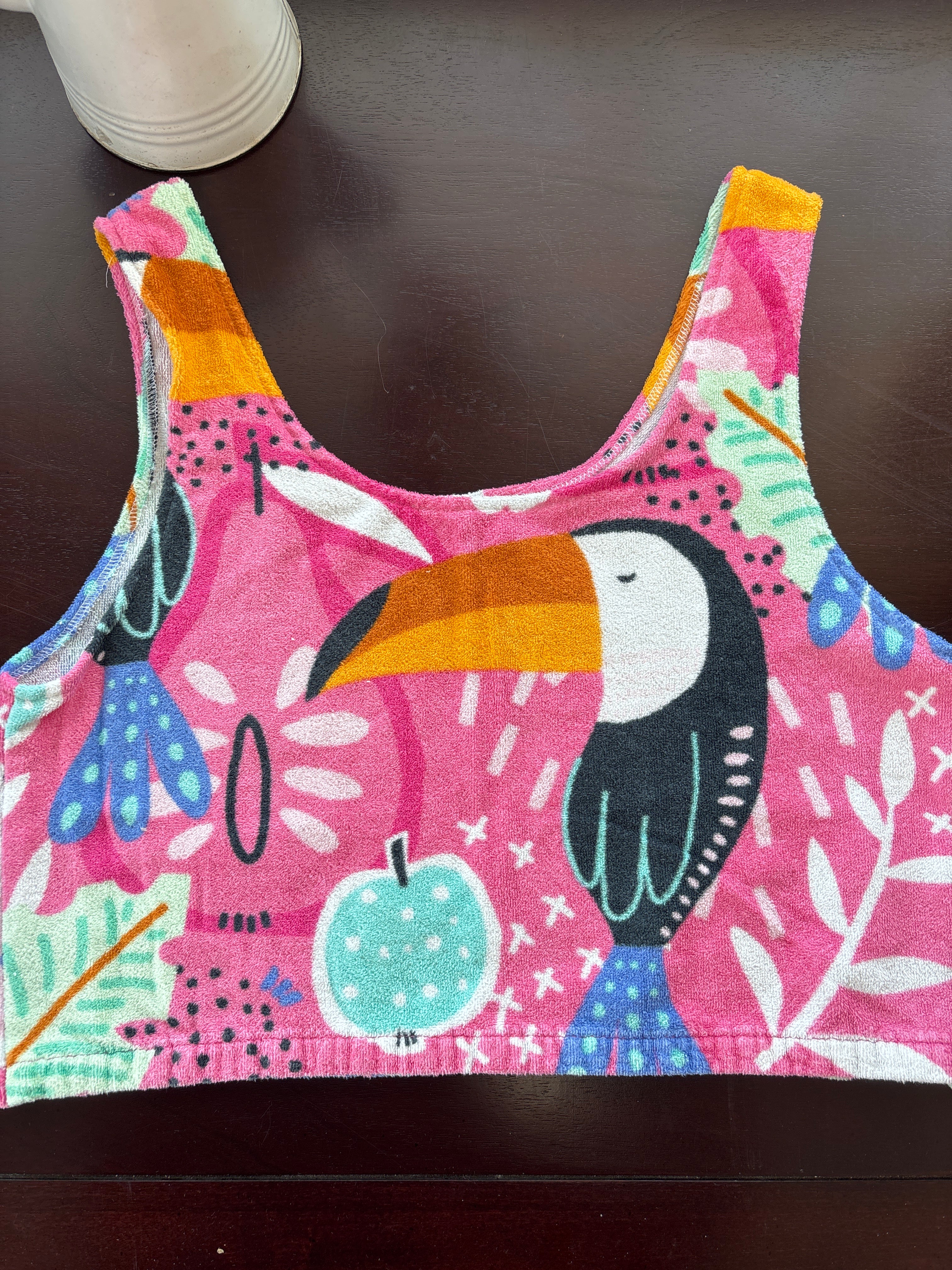 A colorful tank top with a tropical bird and floral print, laid flat on a dark brown surface.