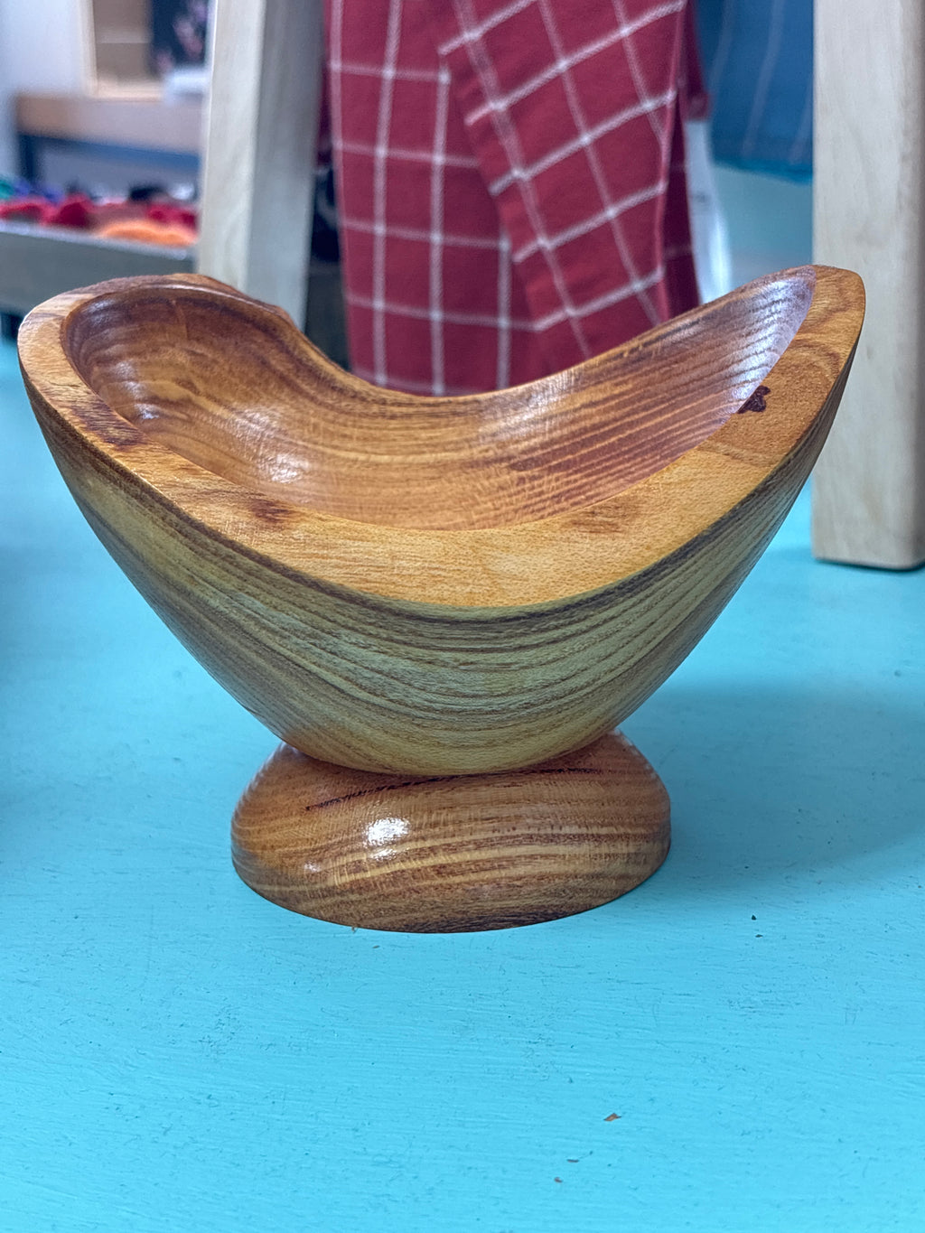 A handcrafted wooden bowl with a matching round pedestal base, displayed on a bright turquoise surface. The background shows a blurred indoor setting with a red and white checkered garment.