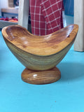 A handcrafted wooden bowl with a matching round pedestal base, displayed on a bright turquoise surface. The background shows a blurred indoor setting with a red and white checkered garment.