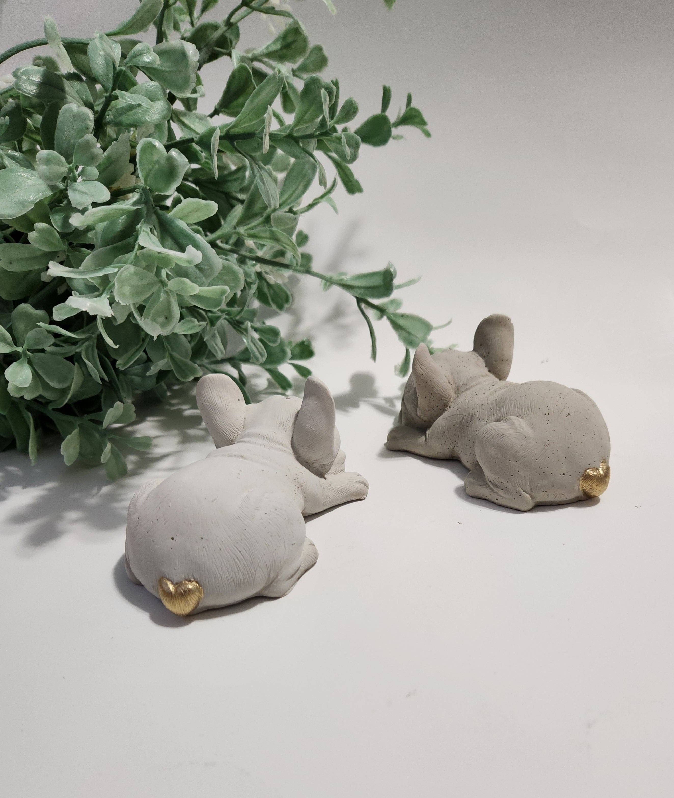 Two small gray clay animals with gold accents on a light surface with green leaves in the background