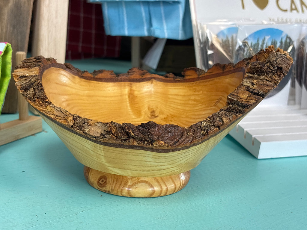 A handcrafted wooden bowl with a natural bark edge and a round pedestal base, shown on a turquoise tabletop with store items blurred in the background.