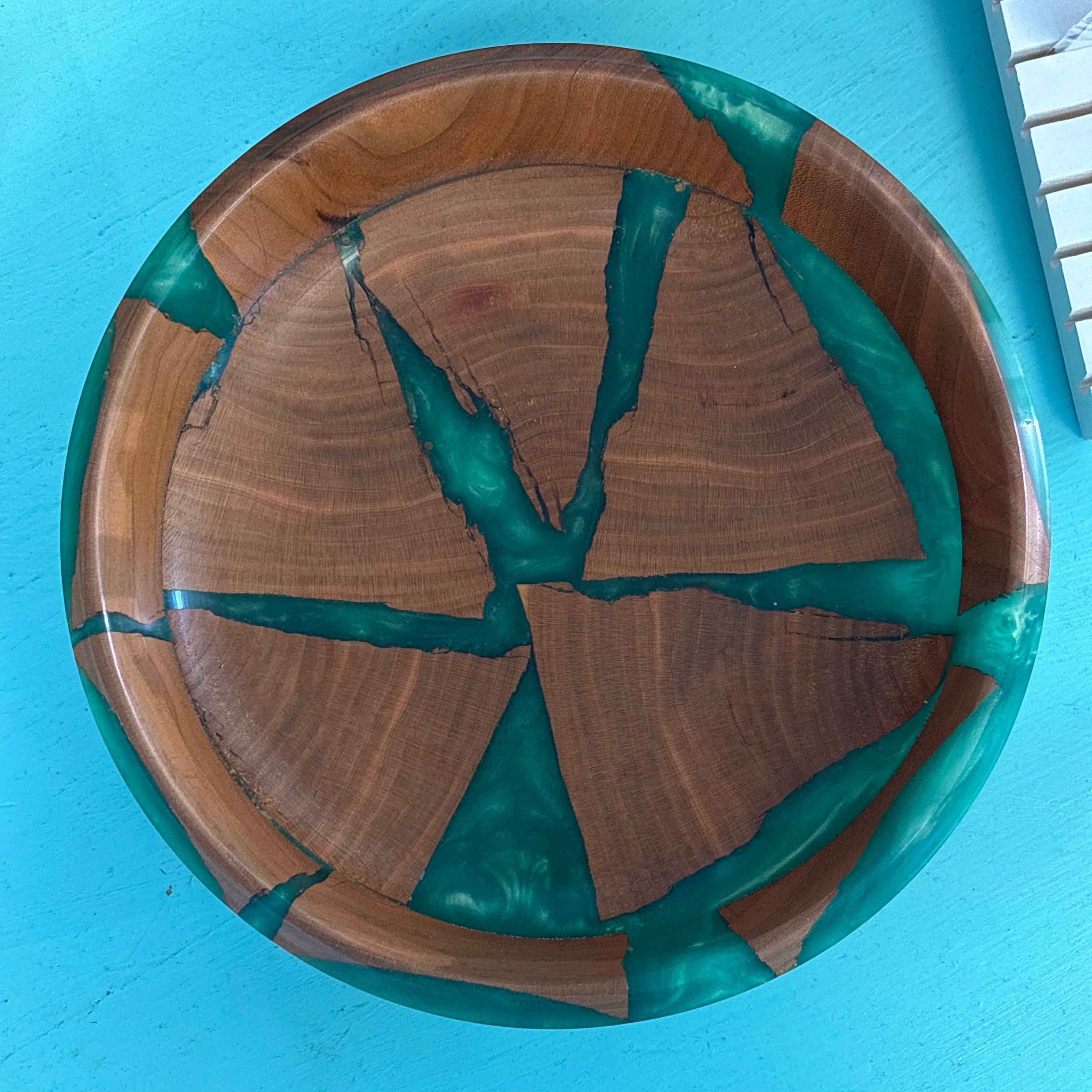 A round wooden bowl with a mosaic pattern of green resin and natural wood grain, photographed from above on a bright turquoise surface.
