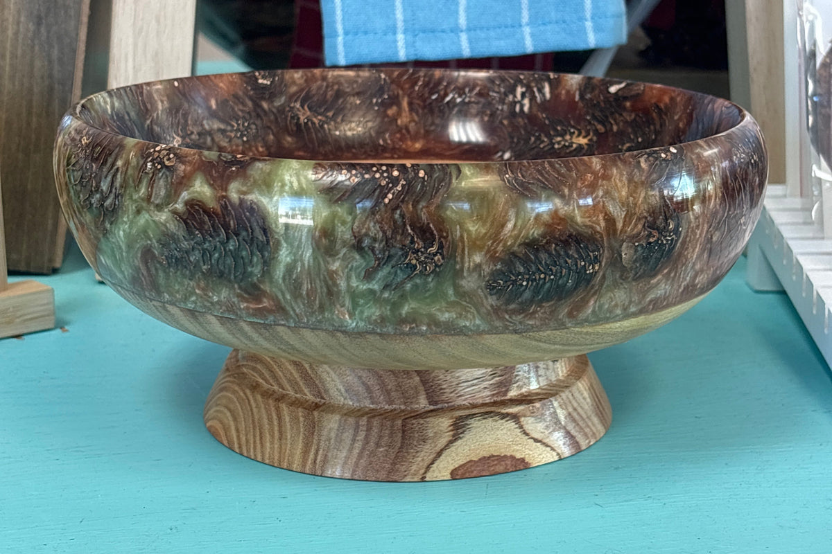 A decorative wooden bowl with a raised pedestal base, featuring a marbled wood grain pattern in brown, green, and black tones. The bowl is displayed on a turquoise surface with rustic wooden furniture and a blue striped towel in the background.