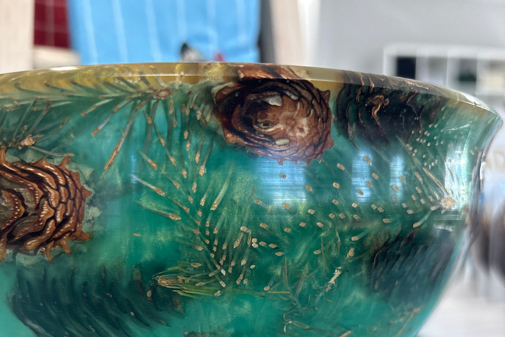 A decorative bowl with a glossy teal and green marbled finish, featuring raised pine needle patterns, pinecone motifs, and a natural wood pedestal base, shown on a turquoise tabletop.