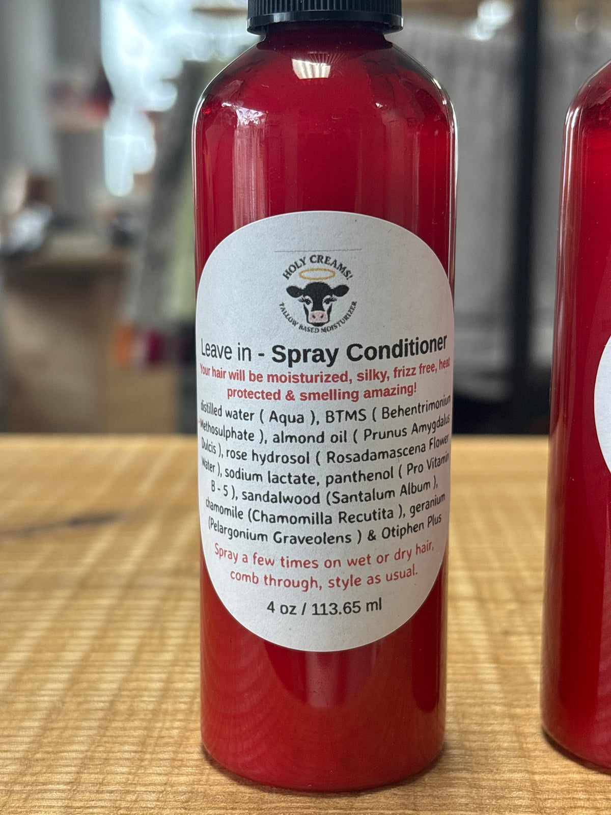 A red plastic bottle of Holy Creams Leave-In Spray Conditioner with a white circular label, sitting on a wooden surface with a blurred background.