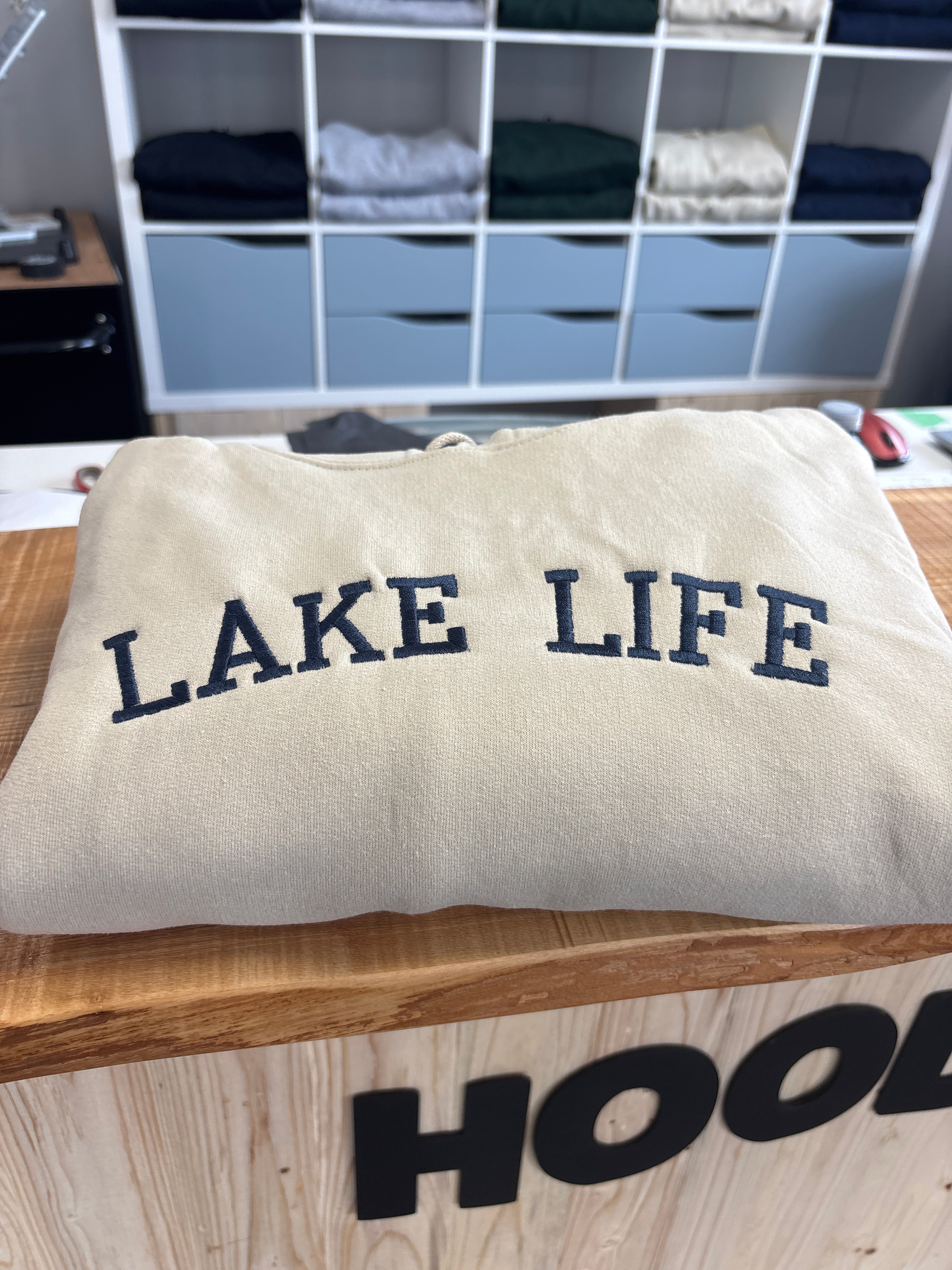 sand hoodie with 'LAKE LIFE' text on a wooden surface with a storage unit in the background.