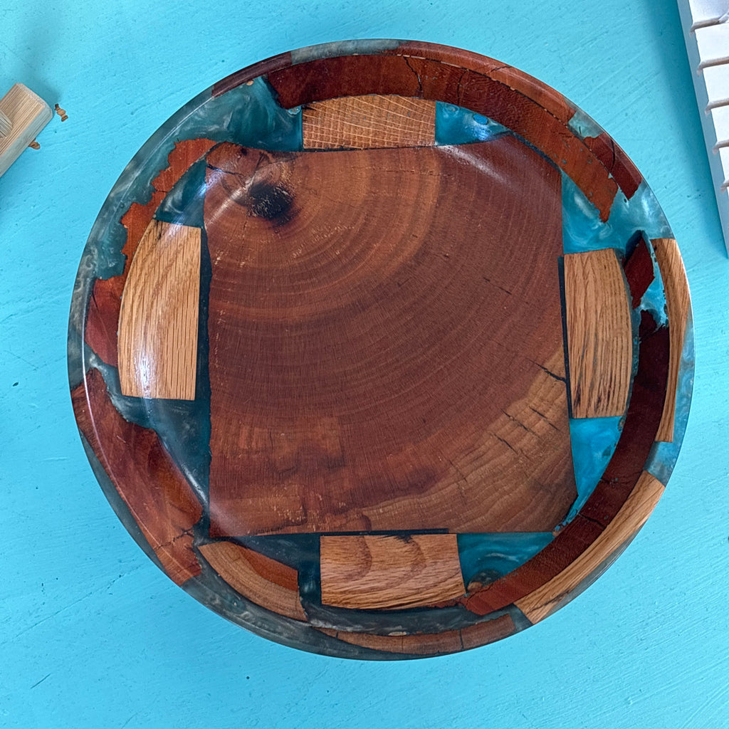 A top-down view of a handmade wooden bowl with a square wood grain center and geometric wood inlay patterns, set against a bright turquoise background.