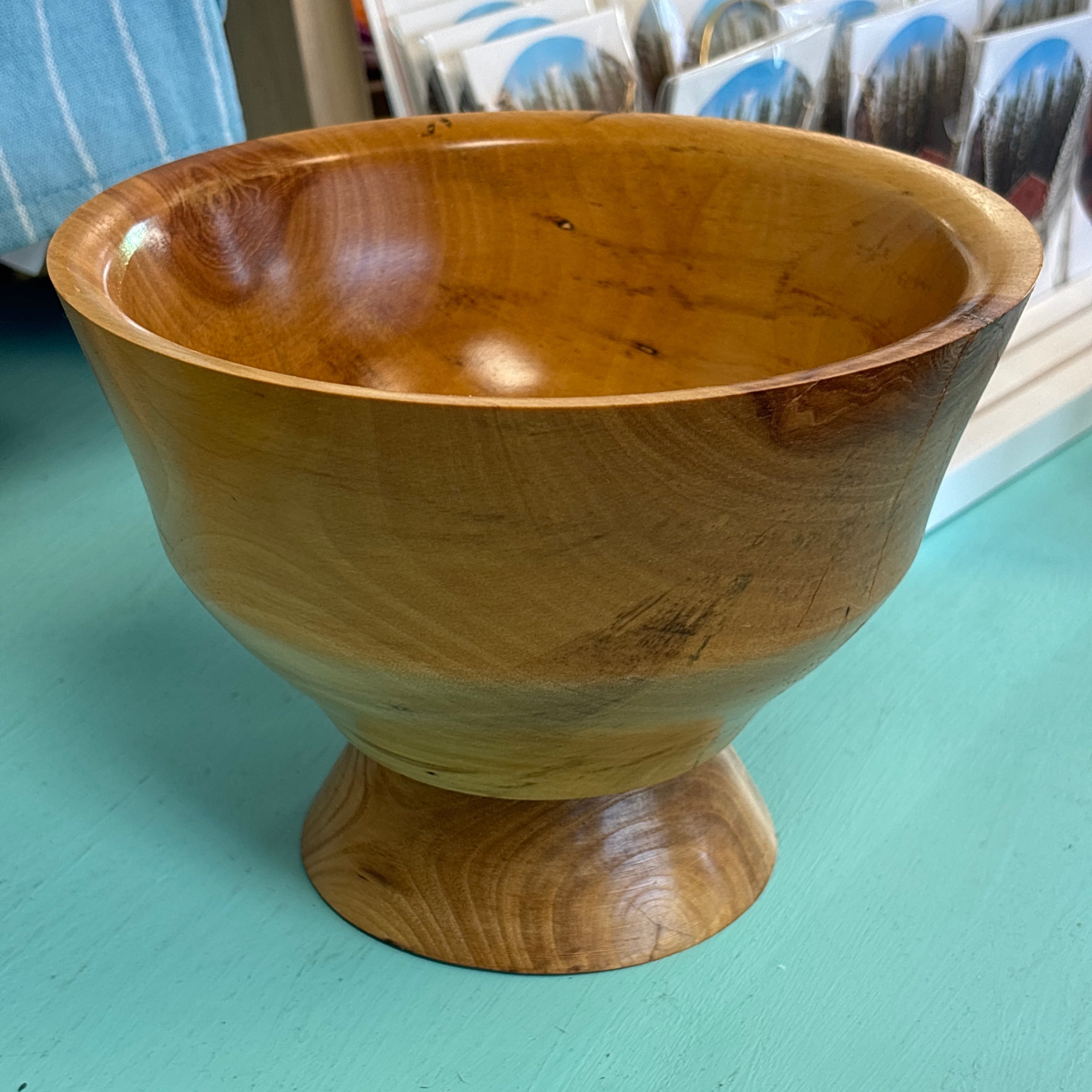 A handcrafted wooden pedestal bowl with a rich warm brown color and visible wood grain, set on a turquoise painted surface with a store display in the background.