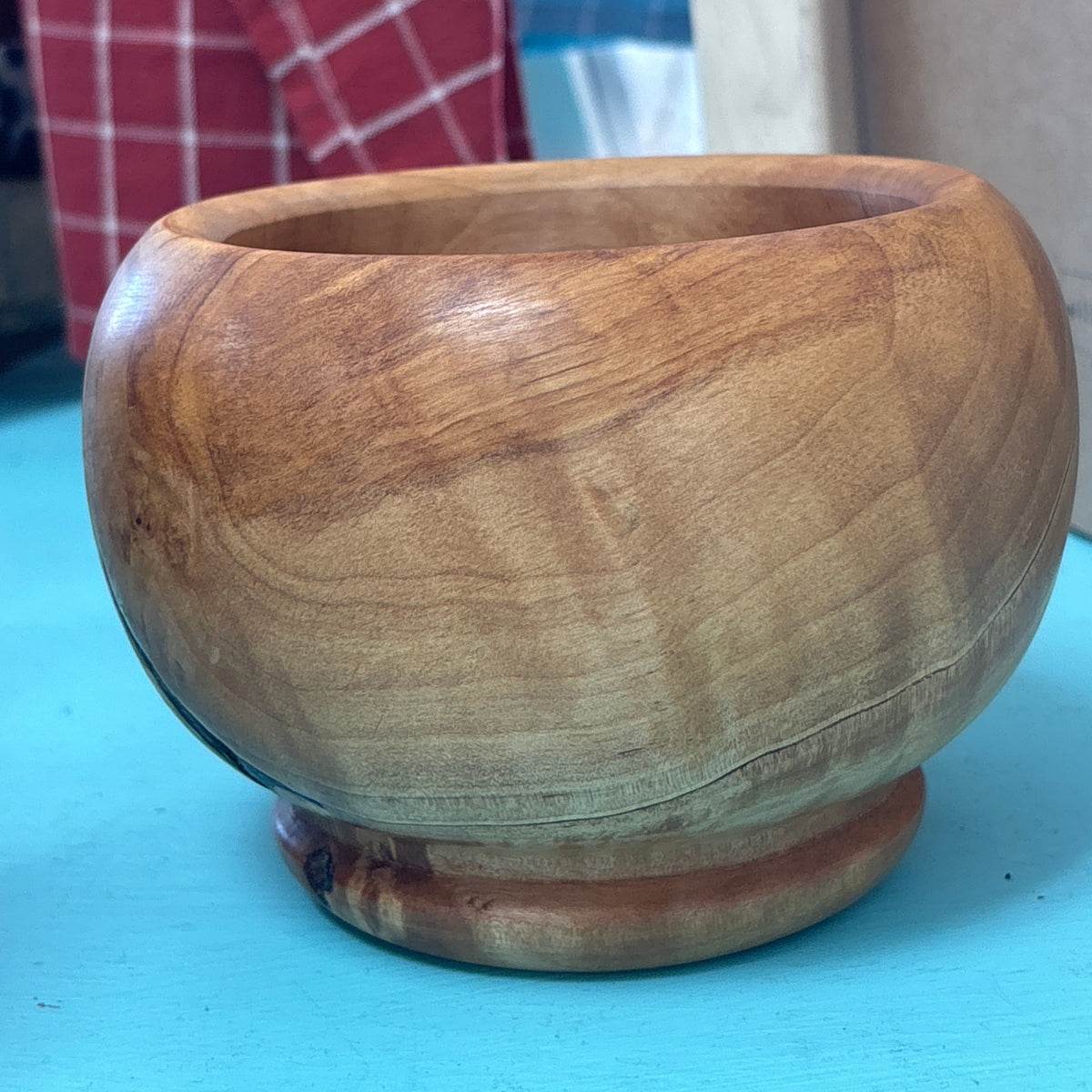 A round wooden bowl with a small pedestal base, made from natural wood with visible grain patterns, sitting on a bright blue surface.