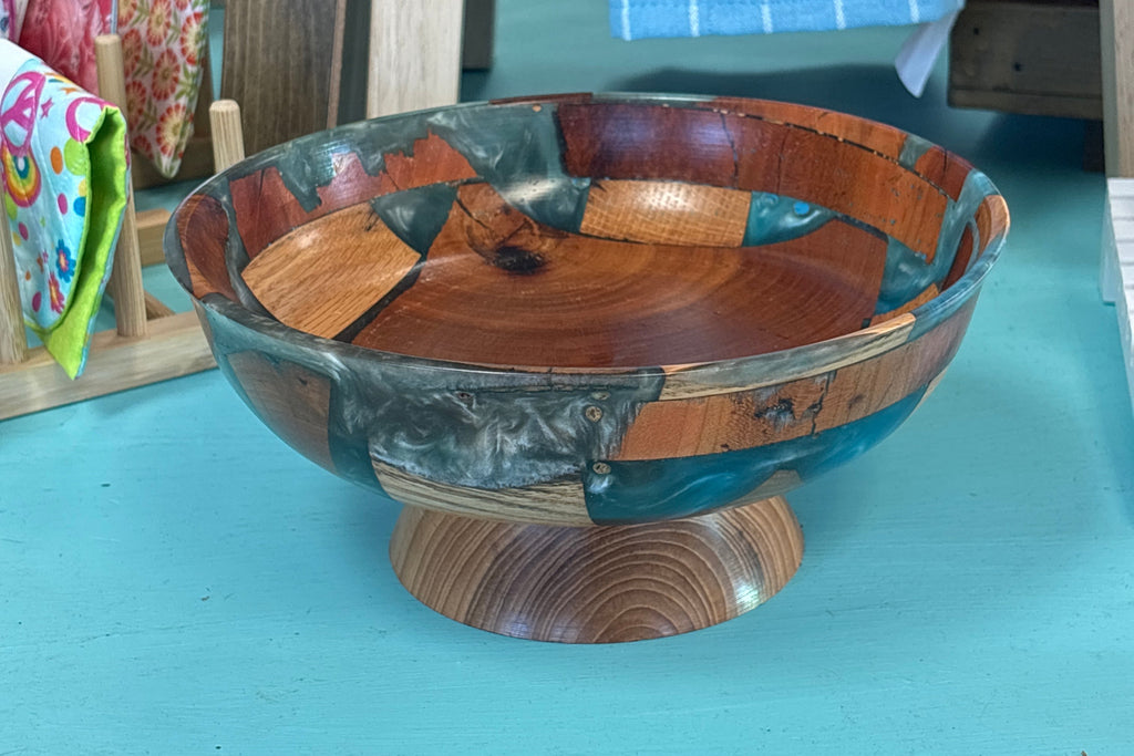 A handmade wooden bowl with a tiered pedestal base, crafted from various wood tones and teal-colored inclusions, displayed on a turquoise table.