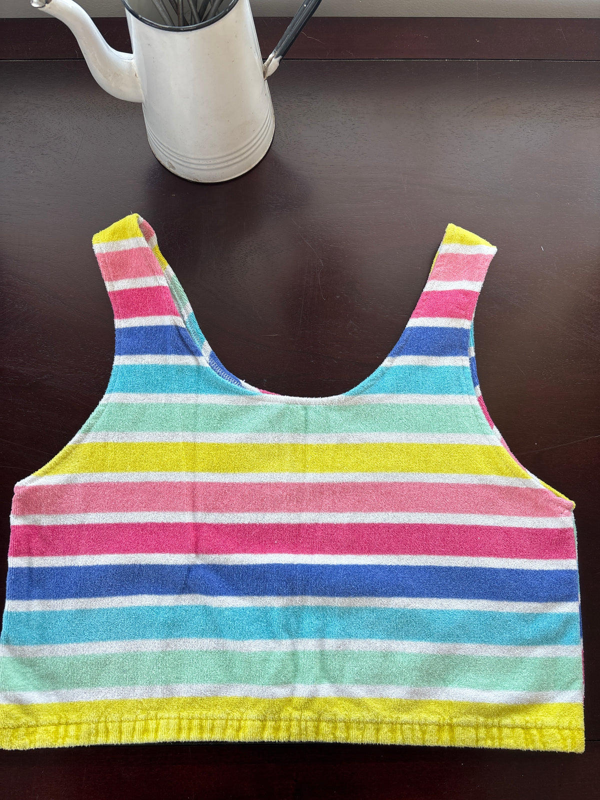 A striped tank top towel laid flat on a dark brown wooden table, featuring vertical stripes of pink, blue, yellow, mint green, and white. 
