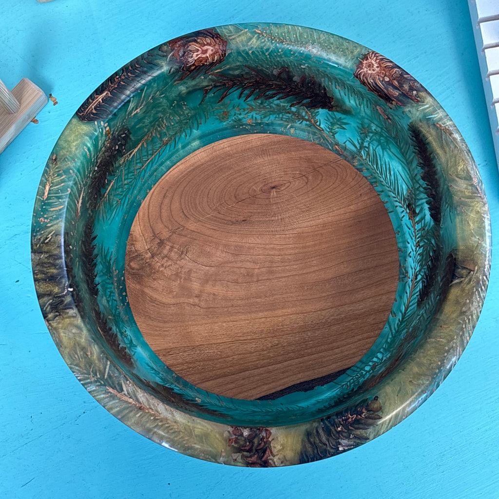 A round wooden bowl with a vibrant turquoise and green resin finish and natural wood grain, photographed from above on a bright blue surface.