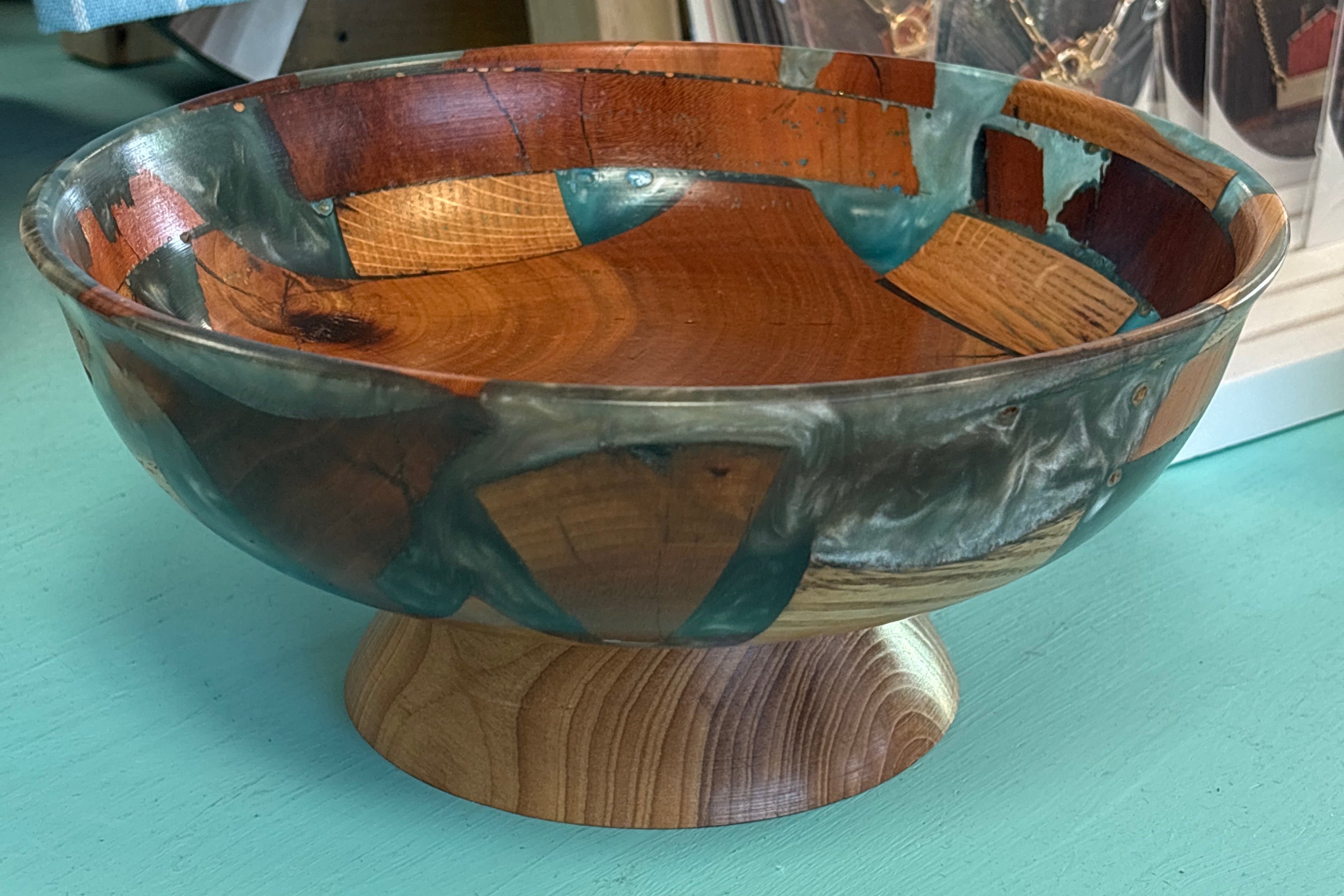 A handcrafted wooden pedestal bowl with a patchwork design of natural wood tones and teal resin, displayed on a turquoise table with a blue striped towel and a 'I ♥ CANADA' sign in the background.
