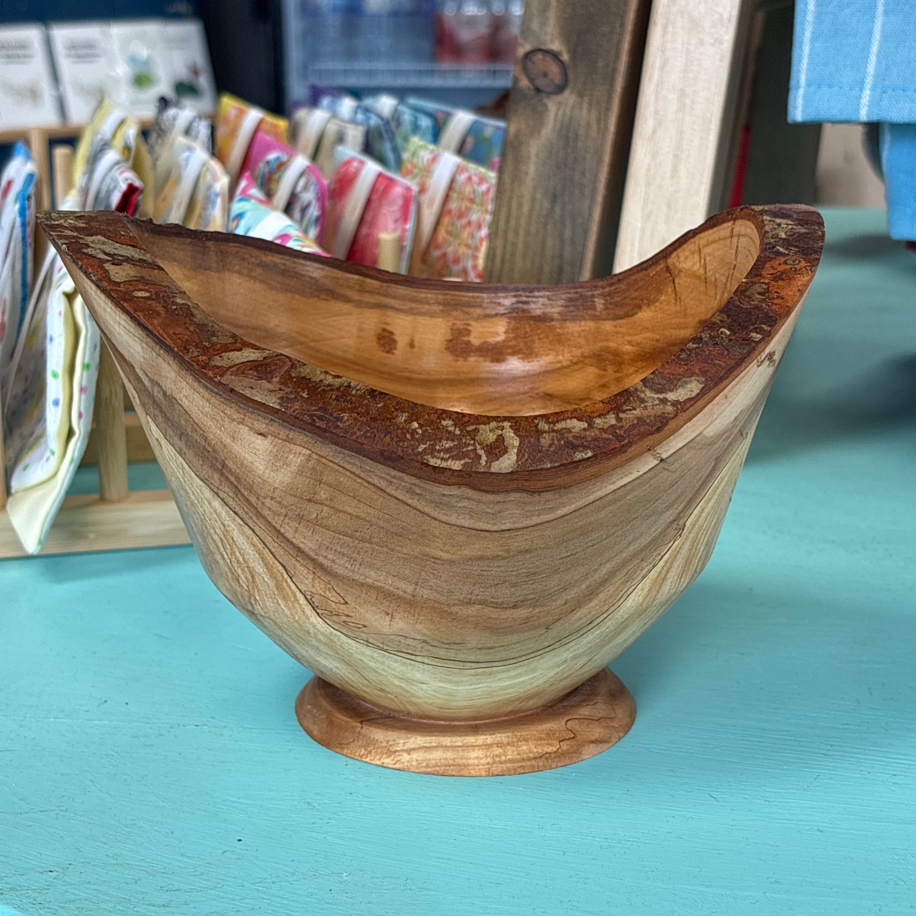 A handcrafted wooden bowl with a natural bark edge and visible wood grain, displayed on a turquoise surface in a shop setting.
