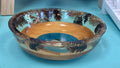 A handcrafted wooden bowl with a colorful tropical landscape design and dark brown pine tree silhouettes, displayed on a turquoise painted surface.