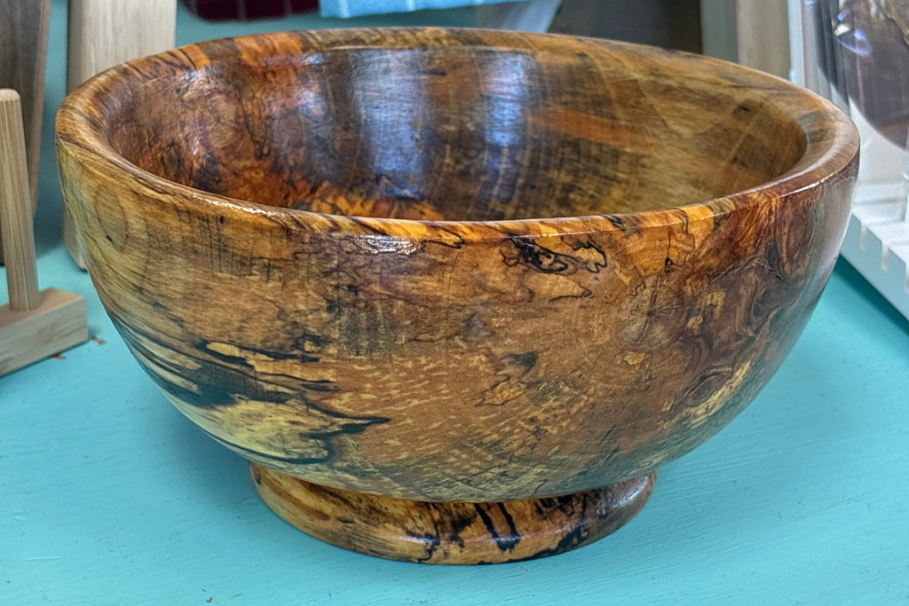 A handcrafted wooden bowl with a rich, swirling natural wood grain pattern in shades of brown and golden yellow, sitting on a turquoise painted surface.