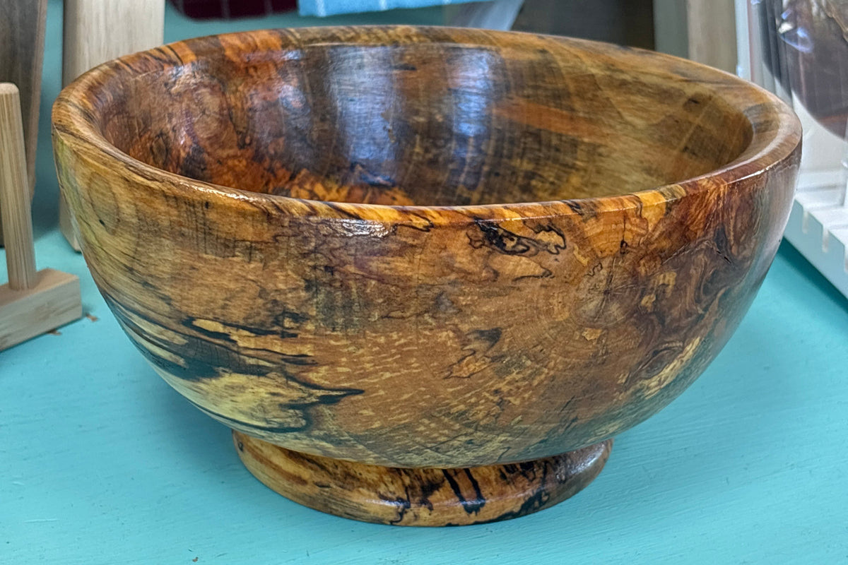 A handcrafted wooden bowl with a rich, swirling natural wood grain pattern in shades of brown and golden yellow, sitting on a turquoise painted surface.