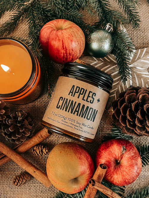 Candle labeled 'Apples + Cinnamon' surrounded by apples, cinnamon sticks, and pinecones on a rustic background.