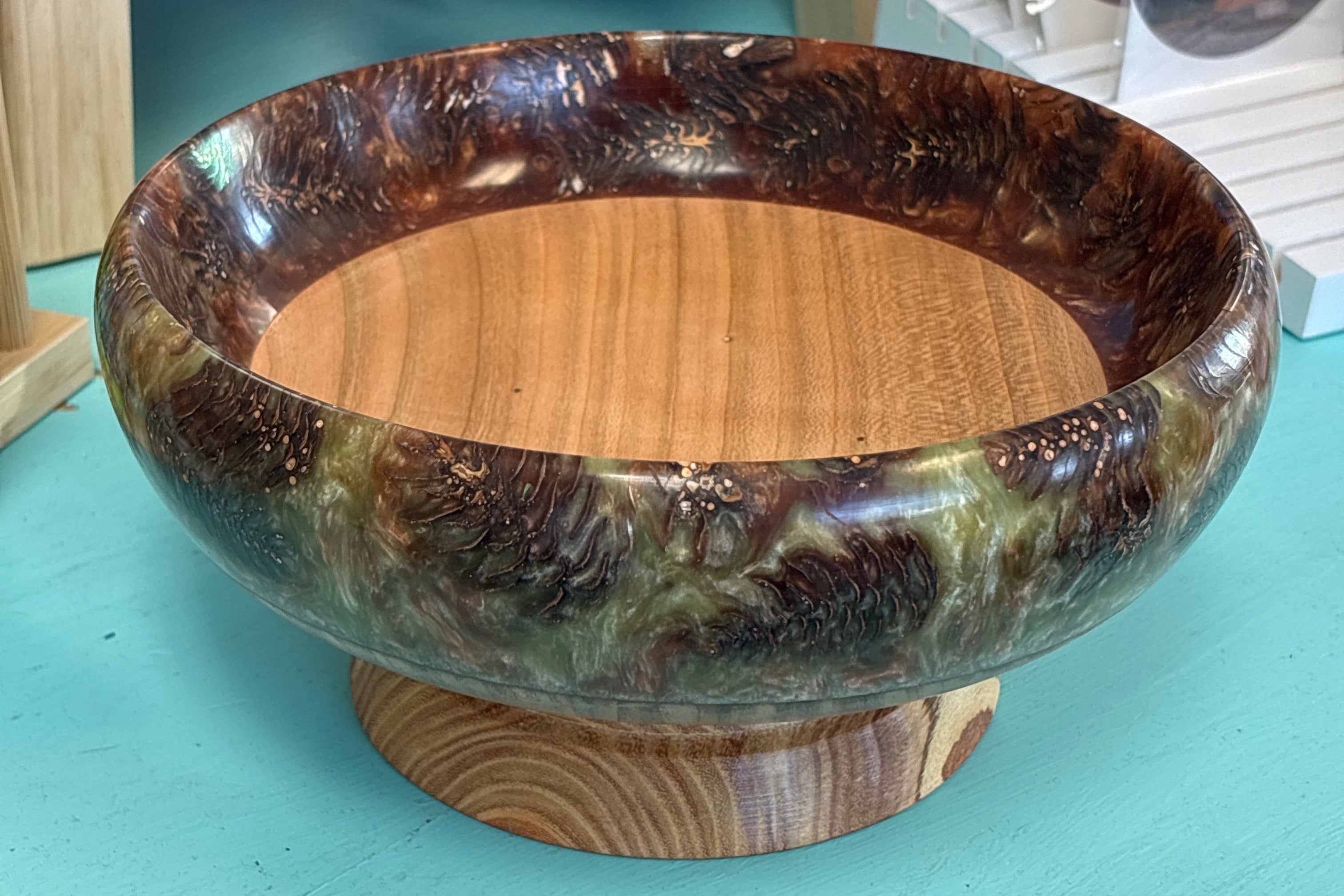 A round wooden bowl with a raised pedestal base, featuring a glossy dark brown and black marbled exterior and a natural light wood interior, displayed on a turquoise table.