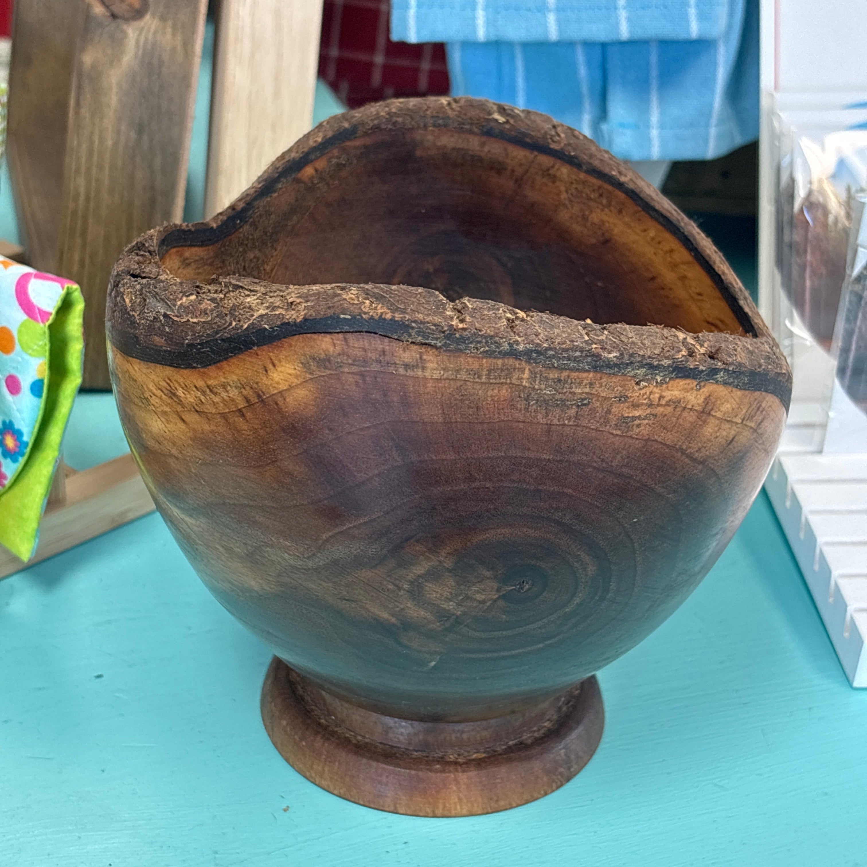 A rustic hand-carved wooden bowl with a natural irregular edge and a small pedestal base, shown on a turquoise surface with a shop or home interior in the background.
