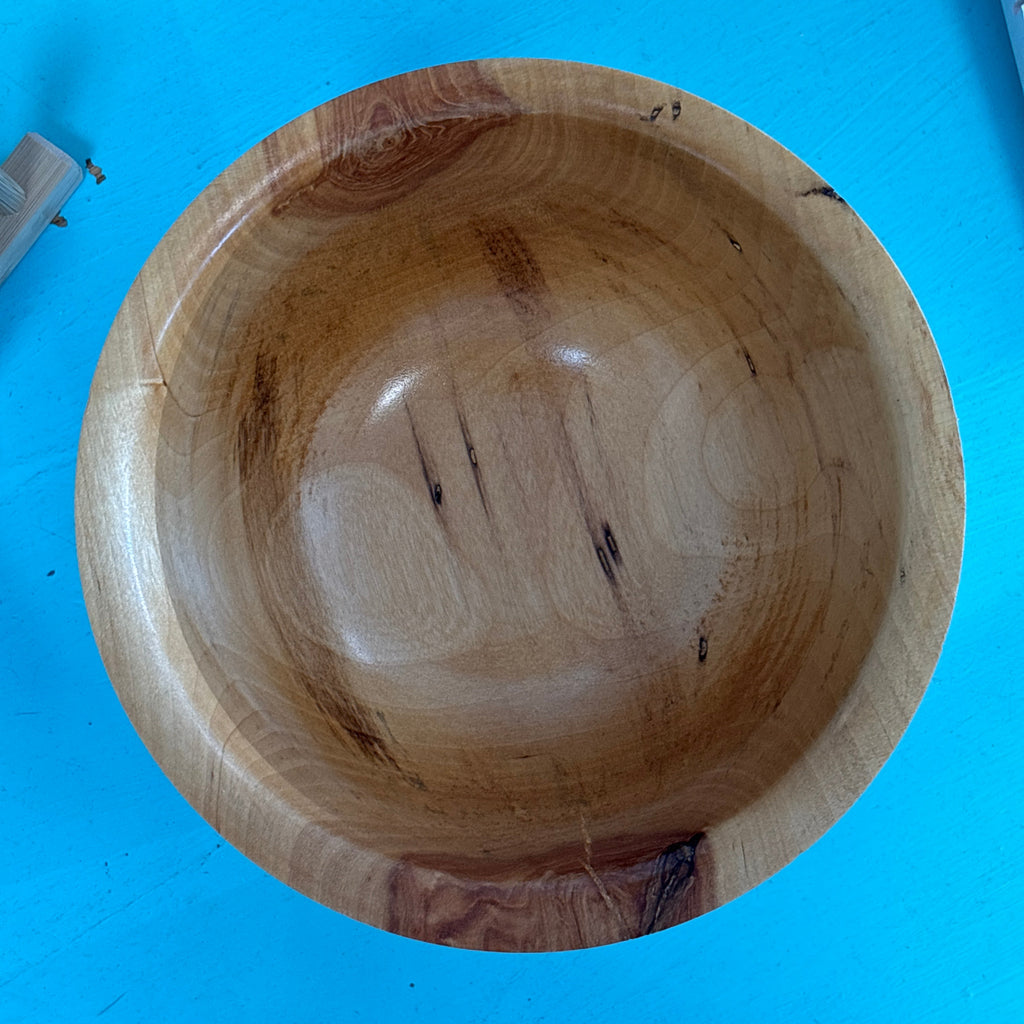 A handcrafted wooden bowl with visible natural grain patterns and dark knots, photographed from a top-down perspective on a bright blue surface.