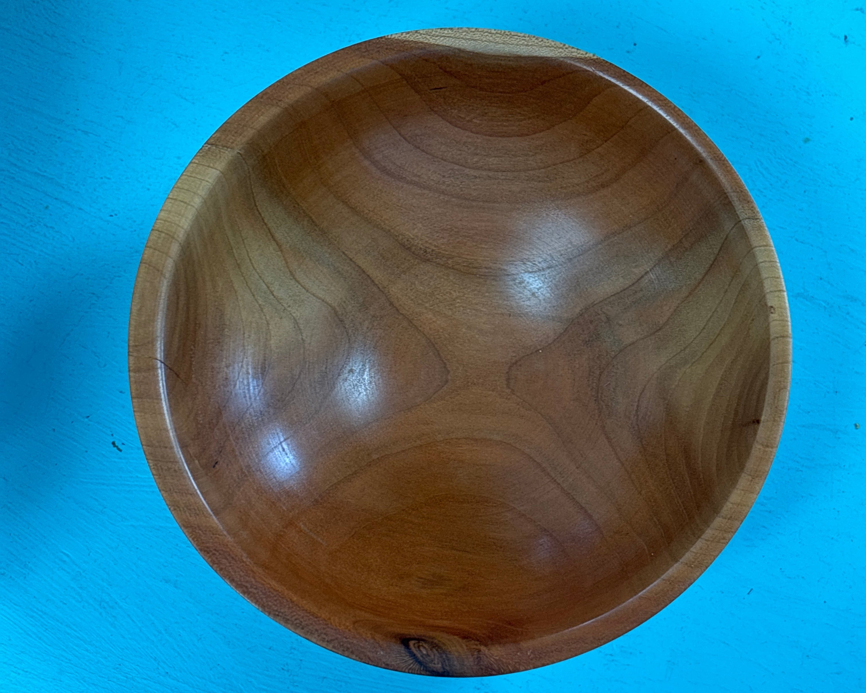 A round wooden bowl with a polished finish, showcasing natural wood grain patterns, photographed from directly above against a bright blue background.