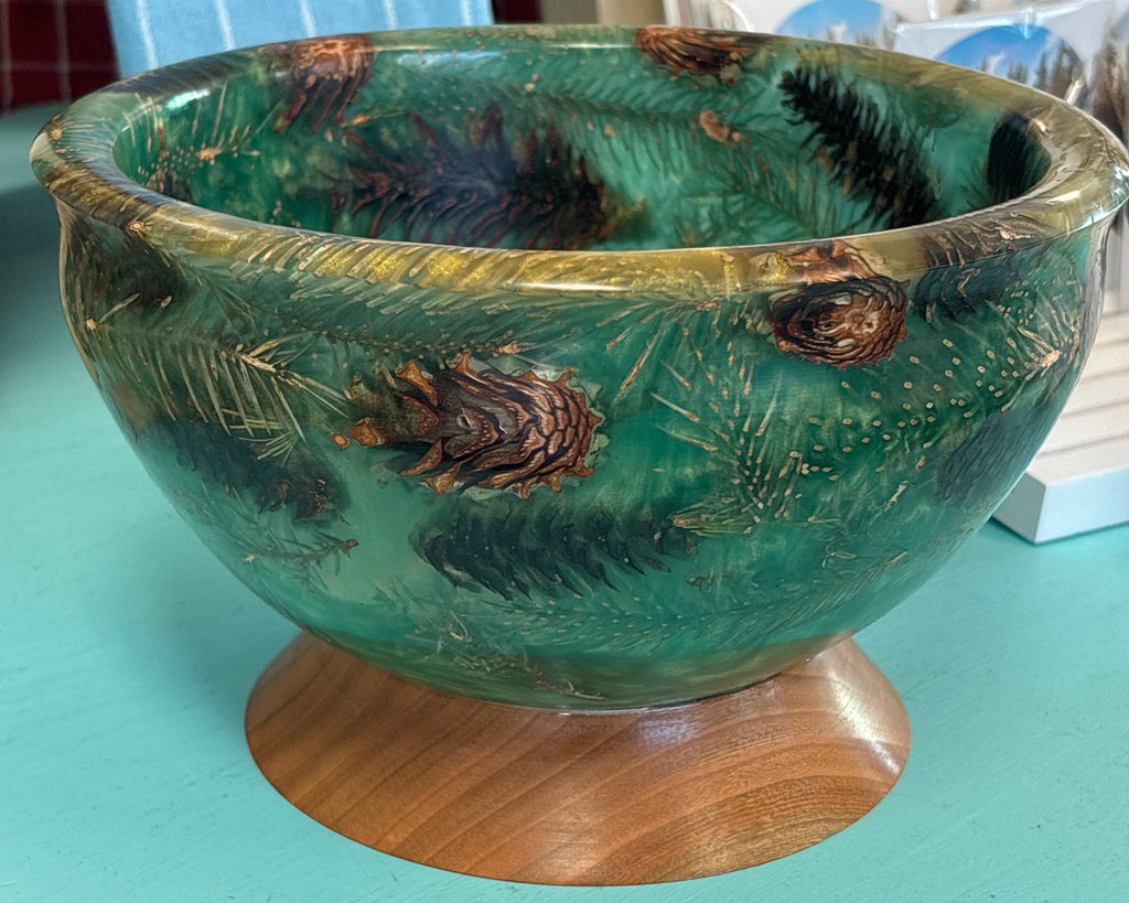 A decorative green glass bowl with pine needle and pinecone motifs, displayed on a round wooden stand against a turquoise tabletop.