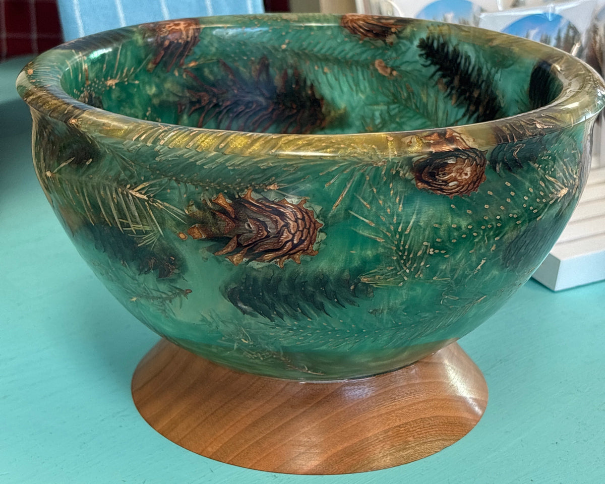 A decorative green glass bowl with pine needle and pinecone motifs, displayed on a round wooden stand against a turquoise tabletop.