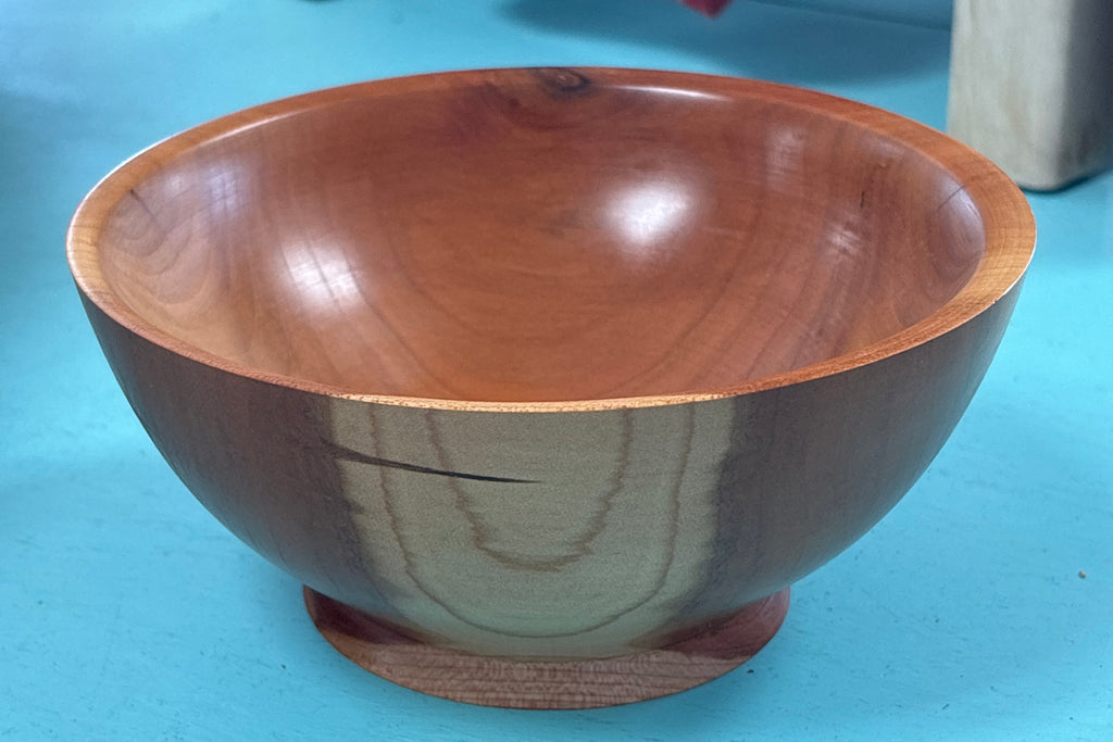 A handcrafted wooden bowl with a glossy finish and a small round wooden base, set on a bright turquoise floor with a blurred background showing store furniture.