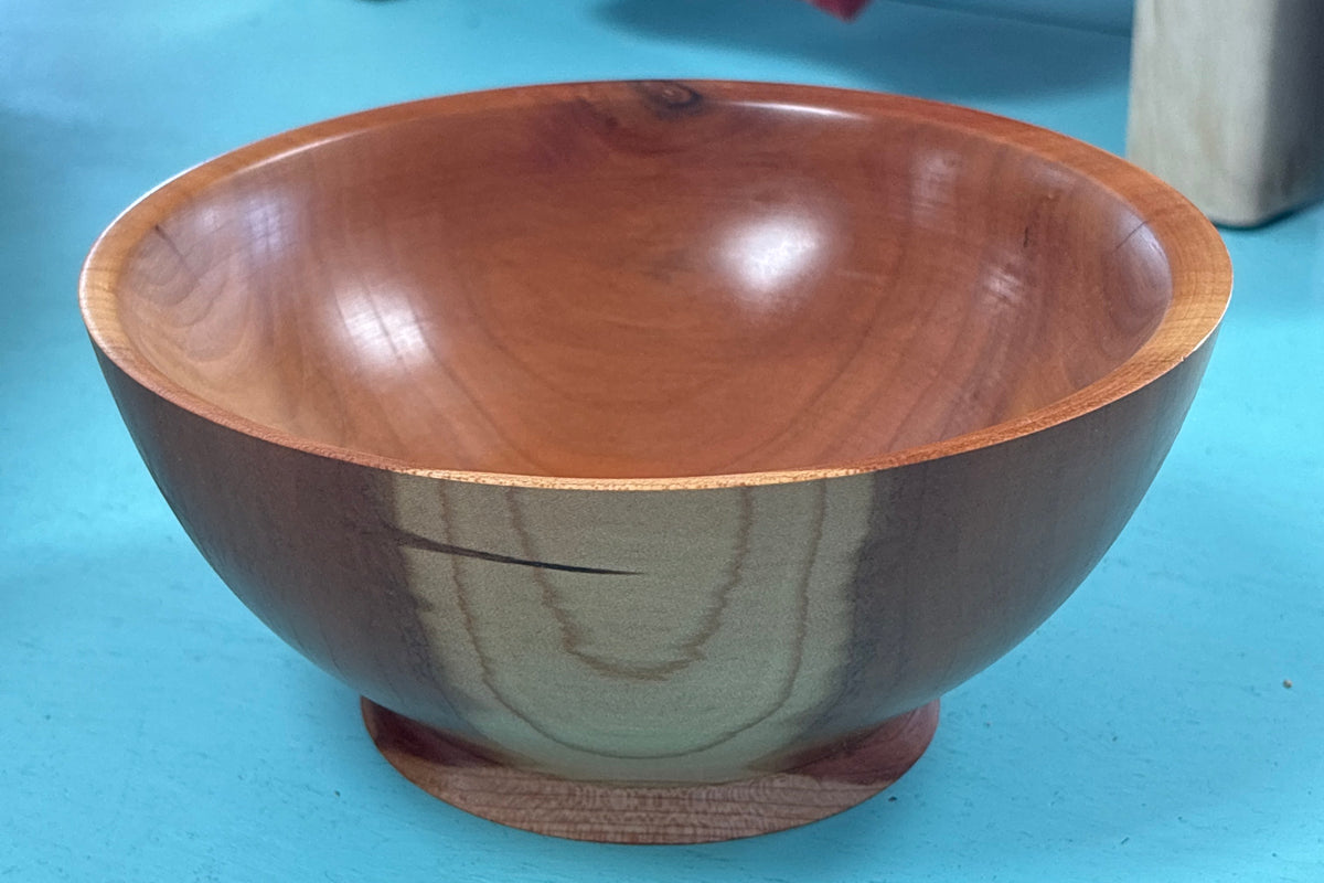 A handcrafted wooden bowl with a glossy finish and a small round wooden base, set on a bright turquoise floor with a blurred background showing store furniture.