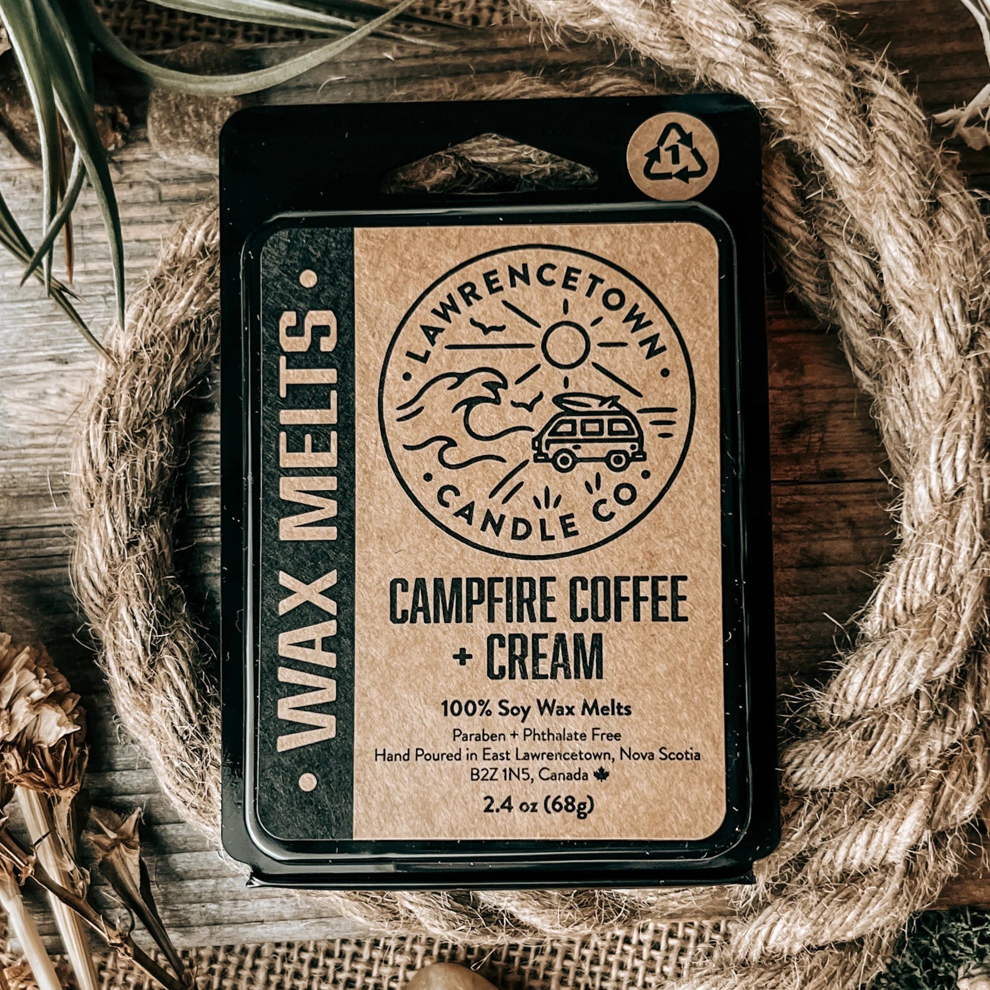 A black rectangular wax melt tin labeled 'Campfire Coffee + Cream' by Lawrence Town Candle Co, styled on a rustic wooden surface with burlap, rope, dried grass, and small stones.