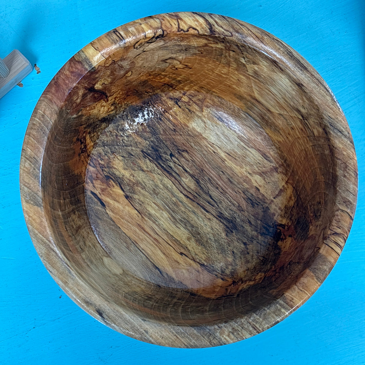 A handcrafted wooden bowl with a glossy finish, showcasing rich natural wood grain patterns in shades of brown, tan, and black, set against a bright blue background.