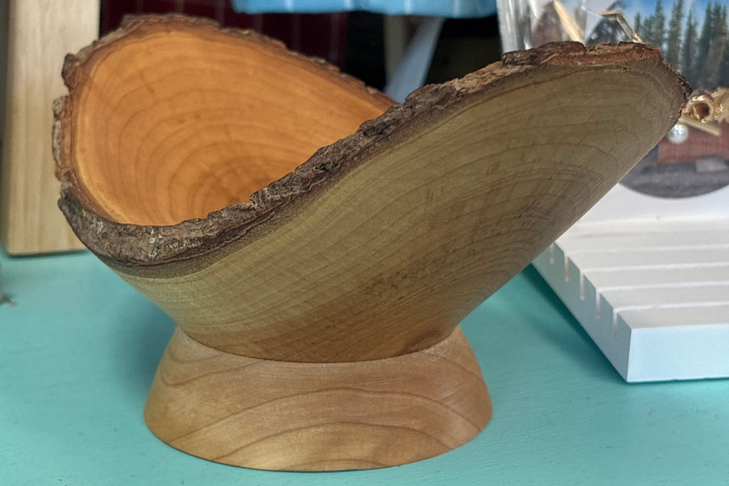 A rustic wooden bowl made from a natural tree slice with bark edges, displayed on a matching wooden pedestal base on a turquoise surface.