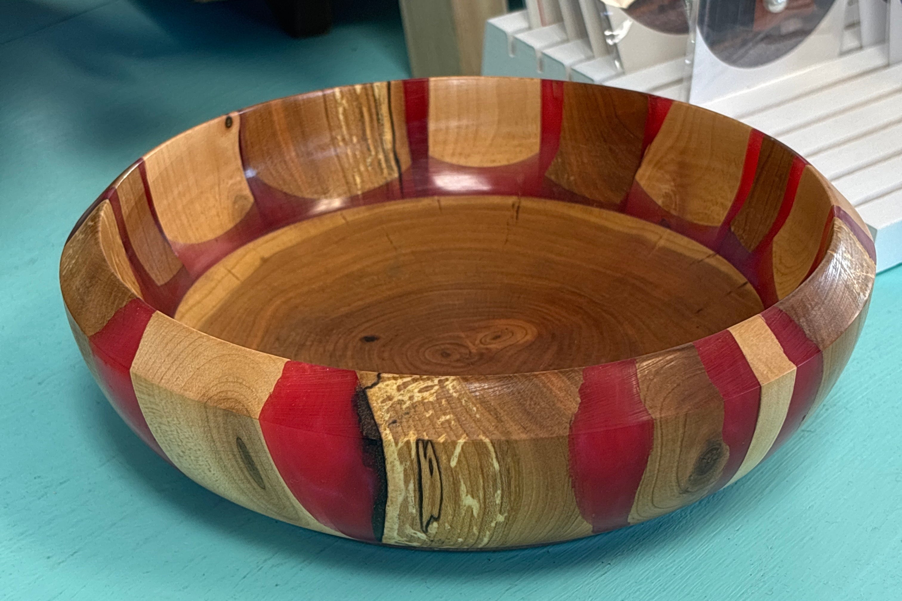 A handcrafted wooden bowl with alternating red, tan, and brown wood stripes, sitting on a turquoise surface.