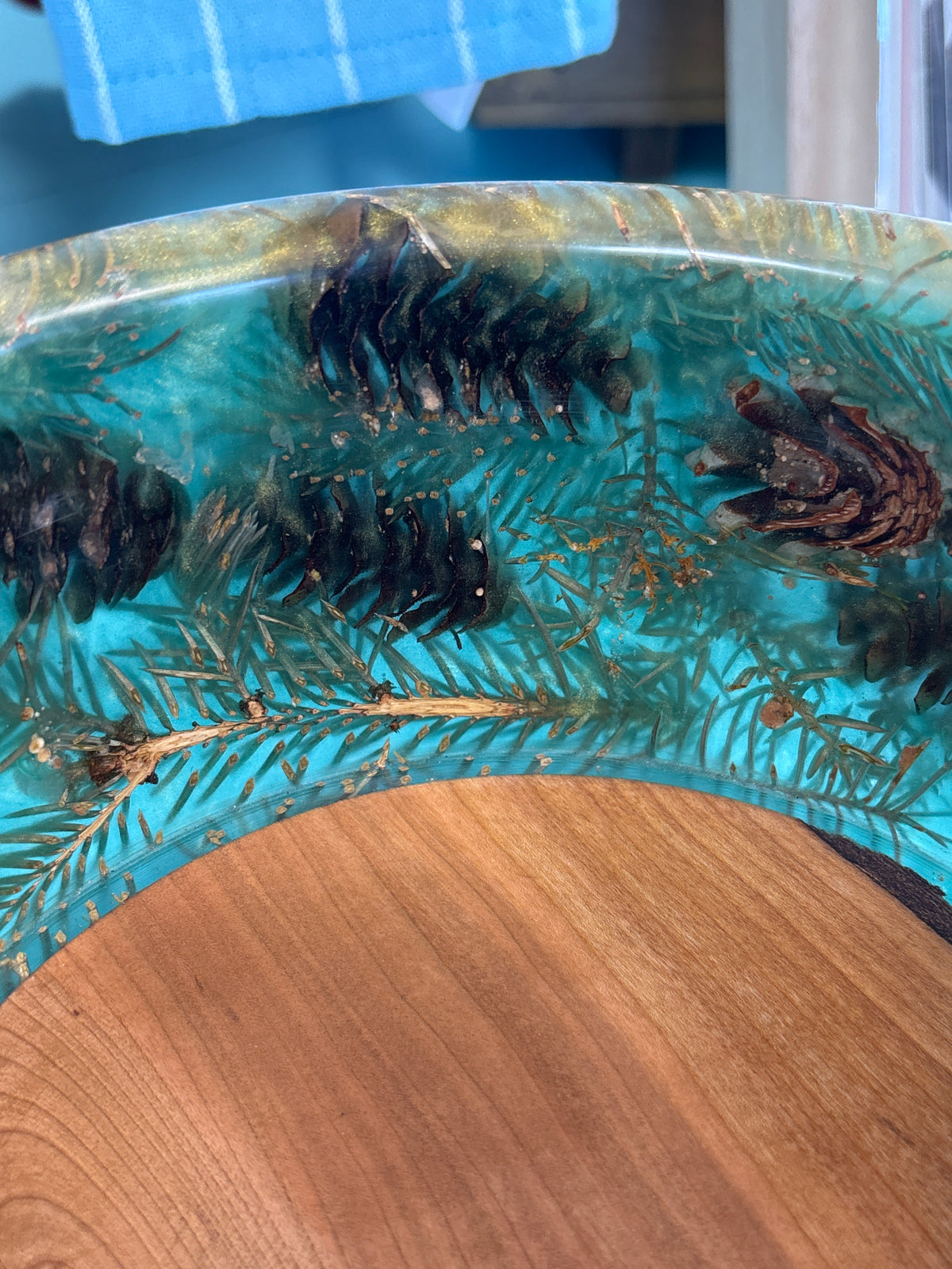 A close-up of a handmade resin tray or bowl featuring turquoise blue resin filled with pine needles, pinecones, and small gold flecks, resting on a wooden surface.
